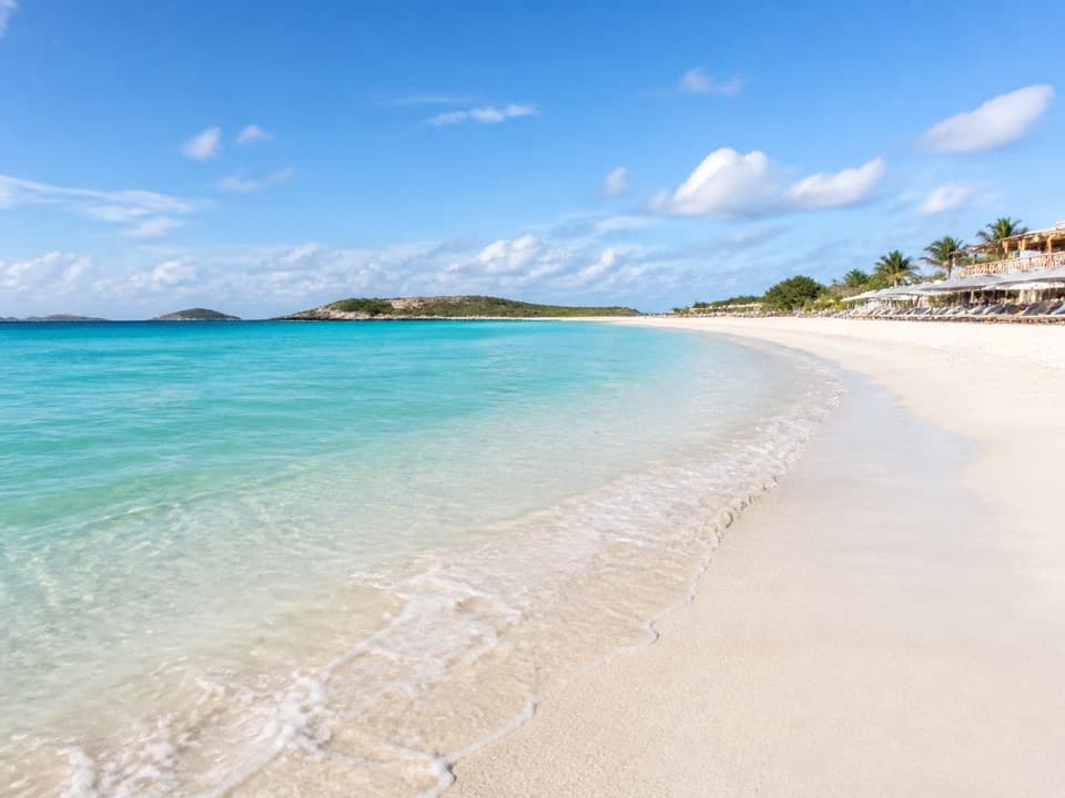 Playa de arena blanca y agua turquesa en Orient Beach, con sombrillas y restaurantes junto a la orilla al fondo.